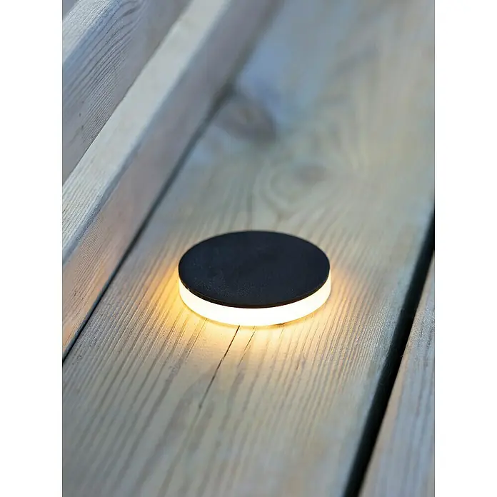 Garden 24 LED-Einbauspot - Warmweiß, Schwarz - Ein flacher, runder, schwarzer LED-Gartenstrahler leuchtet warmweiß auf einer hölzernen Terrassendiele und sorgt für stimmungsvolle Beleuchtung.