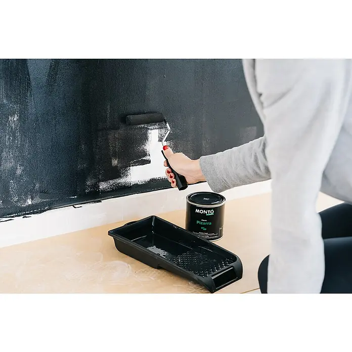 Pintura para efectos decorativos Pizarra - Negro, 500 ml - Persona pintando una pared con pintura Montó Pizarra. También se ven una bandeja y un rodillo de pintura. La pintura es negra y mate.
