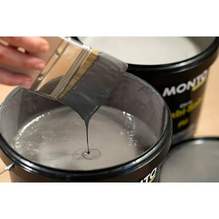 Montó Bricolovers Yeso con efecto de color Wabi Sabi - Gris intenso, Mate - Un pincel se sumerge en un cubo de pintura gris, que gotea del rodillo, con la marca Monto visible al fondo.