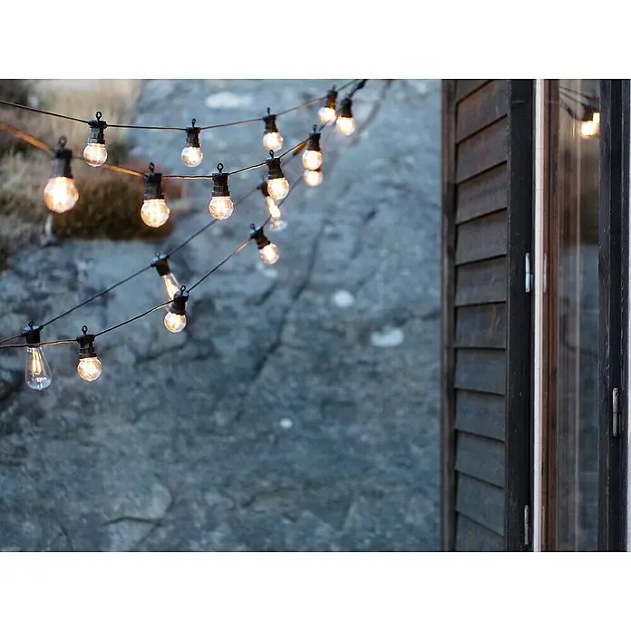 Garden 24 LED-Lichterkette - Außen, 550 cm, Lichtfarbe: Warmweiß, Rund - Lichterkette mit warm leuchtenden Glühbirnen, die eine gemütliche Atmosphäre neben einem rustikalen Holzhaus schaffen.