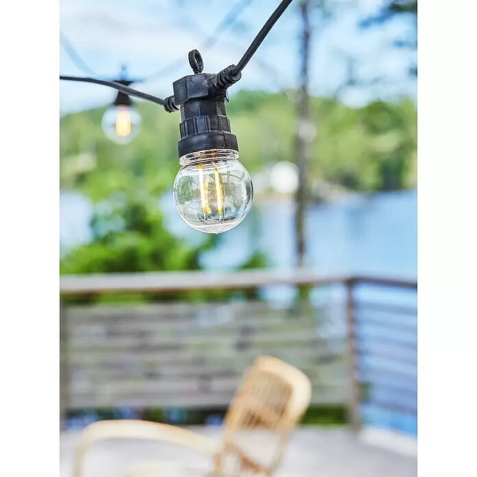Garden 24 LED-Lichterkette - Außen, 550 cm, Lichtfarbe: Warmweiß, Rund - Nahaufnahme einer leuchtenden, klaren LED-Glühbirne einer Lichterkette in schwarzer Fassung, im Freien mit verschwommenem Seeblick.