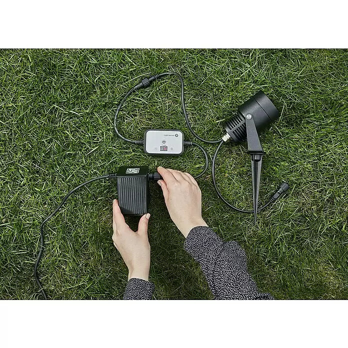Garden 24 Bewegungsmelder Sensor/Timer - Schwarz, L x B x H: 10 x 10 x 3,5 cm - Hände verbinden ein schwarzes Außenstrahlersystem mit grauem Lichtsensor und Timer, bestehend aus schwarzem Transformator und Kabeln.