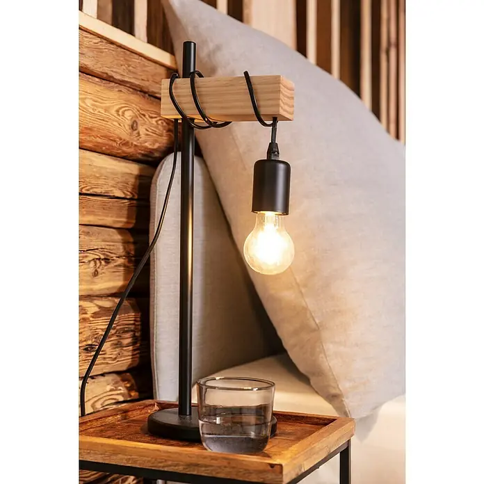 Eglo Townshend Tischleuchte - 10 W, L x B x H: 17,5 x 15,5 x 50 cm, Schwarz, Braun, E27 - Eine moderne Nachttischlampe mit einem schwarzen Gestell und einem Holzbalken, die eine Glühbirne beleuchtet, neben einem Glas Wasser.