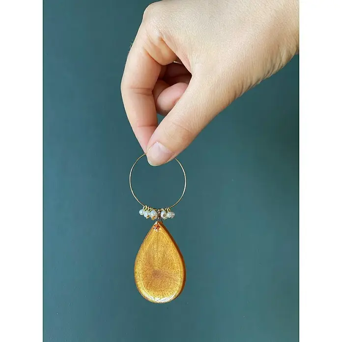 Fleur Molde Pendientes clásicos - Silicona - Primer plano de una mano que sostiene un pendiente de color dorado con un colgante en forma de lágrima sobre un fondo verde azulado.