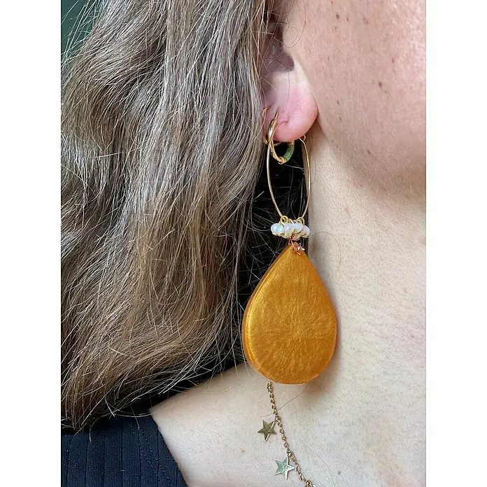 Fleur Molde Pendientes clásicos - Silicona - Primer plano de una oreja con pendientes; un gran pendiente dorado en forma de lágrima cuelga de un aro dorado, adornado con cuentas y colgantes de estrella.