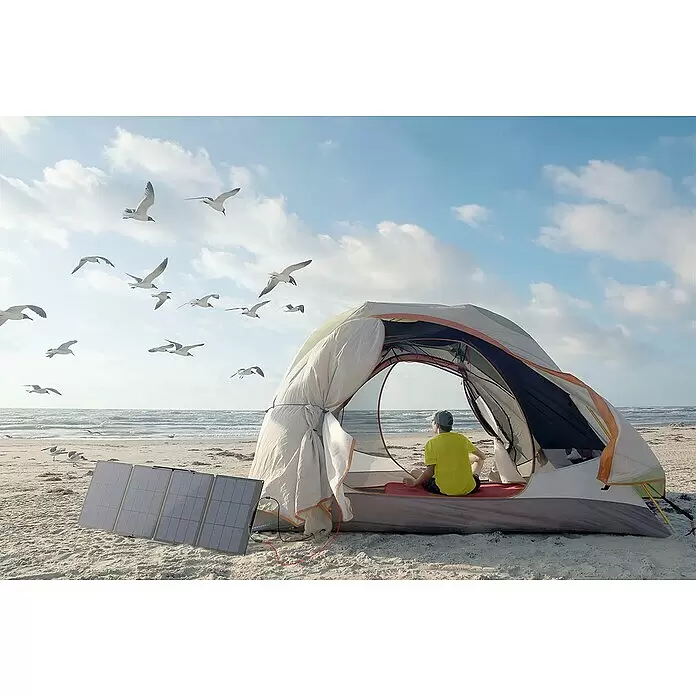 EcoFlow Panel solar Plegable - L x An x Al: 178,5 x 42 x 2,5 cm, 110 W - Panel solar negro plegable en una playa de arena, conectado a una tienda con una persona sentada, mostrando el uso de energía al aire libre.