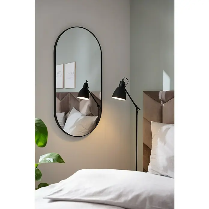 Rahmenspiegel Malva - 60 x 120 cm, Schwarz - Ovaler Spiegel mit schwarzem Rahmen hängt an der Wand und reflektiert das Schlafzimmer mit Lampe und Bett.