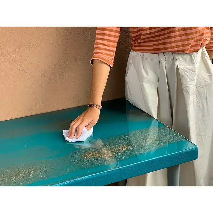 Fleur Purpurina Glitter - Champagne, 90 g, Brillante - Una persona limpia una mesa con una superficie brillante de color teal con detalles dorados, destacando su diseño único.