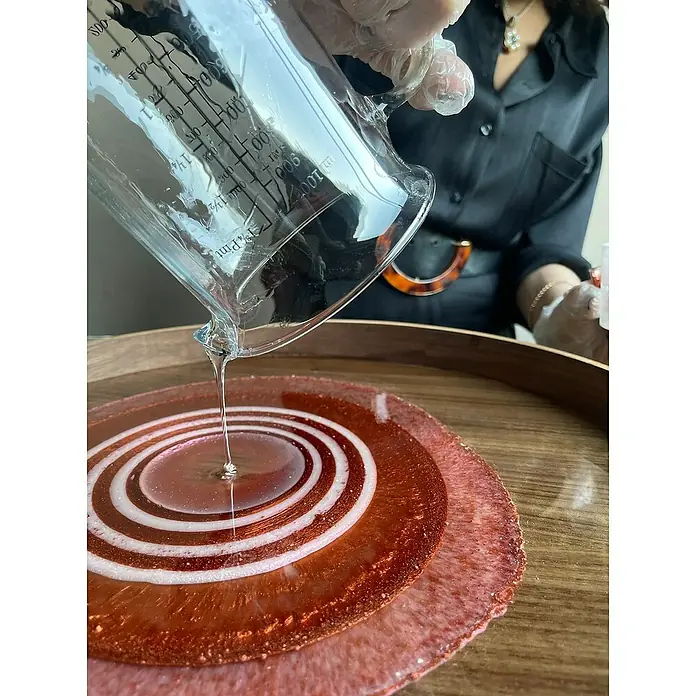 Fleur Resina epoxi Super Transparente - 950 ml, Transparente - Un artesano vierte resina epoxi sobre una obra de arte de resina roja con espirales blancas, que se encuentra sobre una mesa de madera.