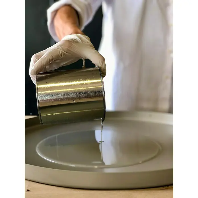 Fleur Resina epoxi Super Transparente - 950 ml, Transparente - Un recipiente metálico vierte un líquido transparente en un plato gris, con una mano enguantada y una bata blanca que complementan la escena.