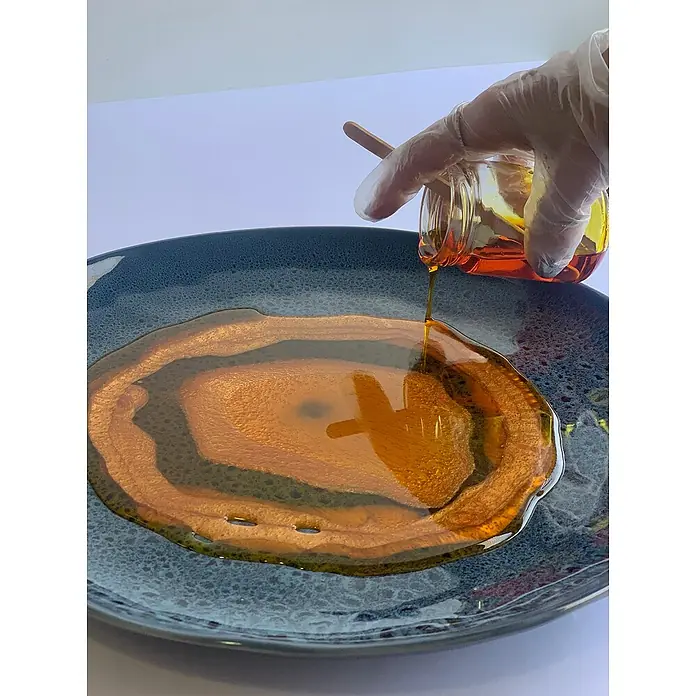 Fleur Resina epoxi Super Transparente - 950 ml, Transparente - Primer plano de un plato azul con resina dorada. Una mano enguantada vierte resina de un tarro sobre un palo de madera.