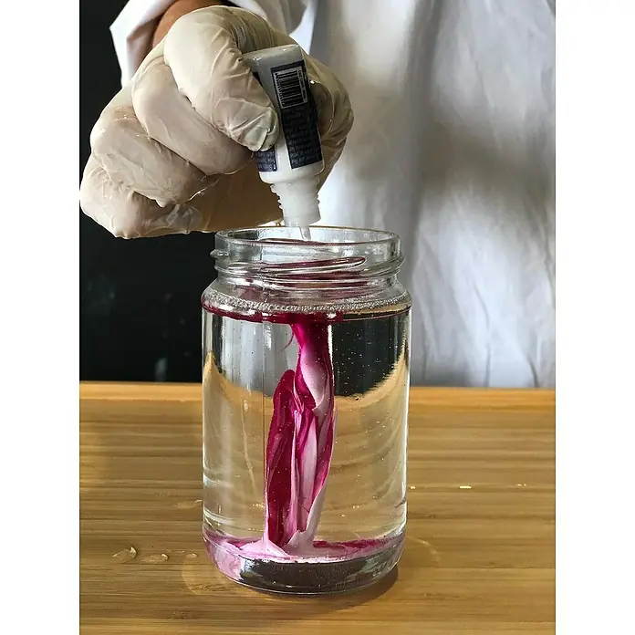 Fleur Colorante líquido para resina - Magenta, 10 ml - Una persona vertiendo colorante rojo en un frasco de agua, con el colorante creando un movimiento de remolino dentro del agua.
