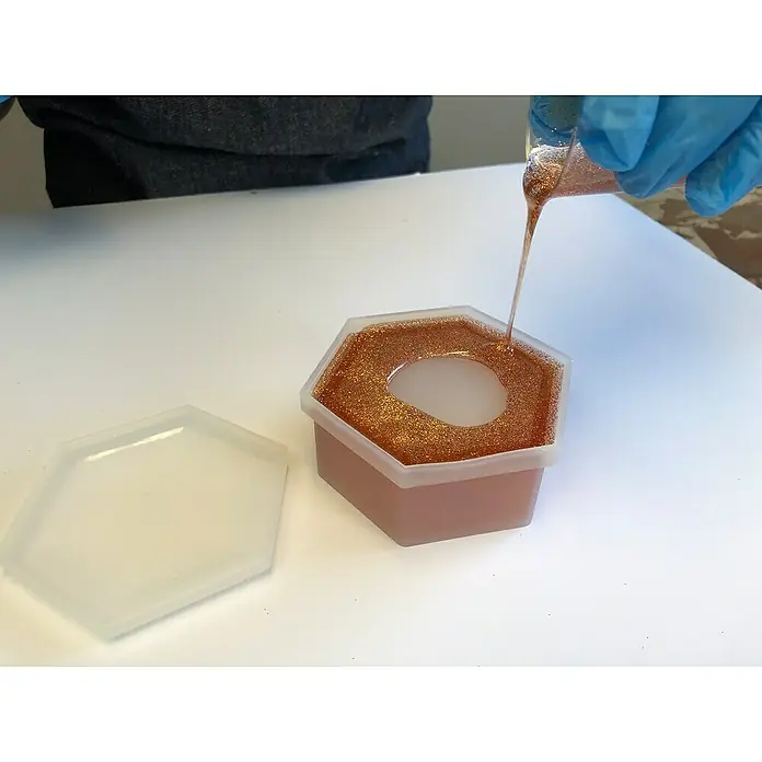 Fleur Molde Joyero - Silicona - Se está vertiendo resina en un joyero hexagonal de silicona con tapa. La caja está llena de copos de brillantina dorada y destellos.