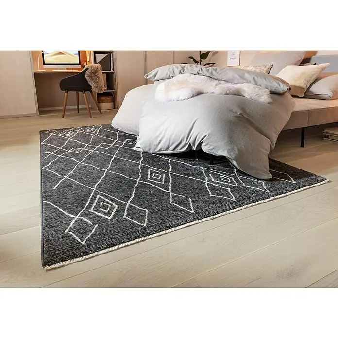 Kurzflorteppich AgadirGraphit, 230 x 160 cm, 100% Polypropylen (Flor) - Ein grauer Teppich mit geometrischem Muster liegt unter einem Bett. Der Teppich hat Fransen und ein modernes Design.
