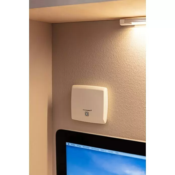 Homematic IP Steuerzentrale Access Point HmIP-HAP - Weiß, 11,8 x 10,4 x 2,6 cm, Funkfrequenz: 868 MHz - 869,4 MHz - Weißes Homematic IP Smart-Home-Gerät, an einer texturierten Wand montiert, beleuchtet von einer Deckenleuchte, neben einem Computerbildschirm.