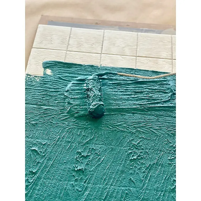 Fleur Kit Microcemento - Greige, 2 kg - Primer plano de la aplicación de pintura con un rodillo, creando una superficie turquesa texturizada sobre un panel con azulejos claros.