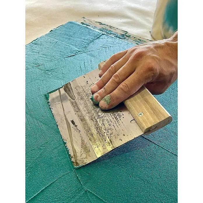 Fleur Kit Microcemento - Grey White, 8 kg - Aplicación de pintura con espátula: Una mano extiende pintura azul sobre un lienzo con una espátula. Distribución precisa del color.