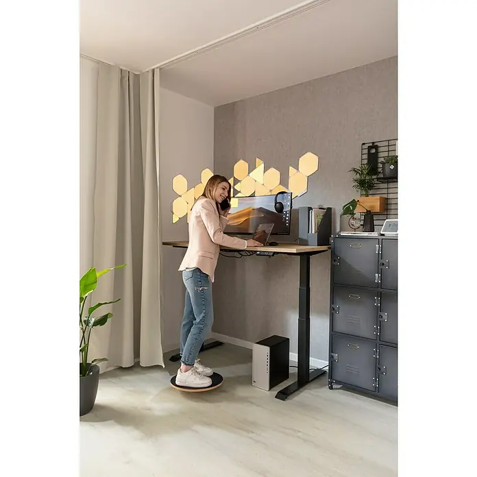 Rasch LOFT Vliestapete Uni - Grau, Betonoptik, 10,05 x 0,53 m - Eine Frau telefoniert an einem höhenverstellbaren Stehtisch mit Laptop, Monitor und Lichtpaneelen an der Wand, während sie auf einem Balance Board steht.