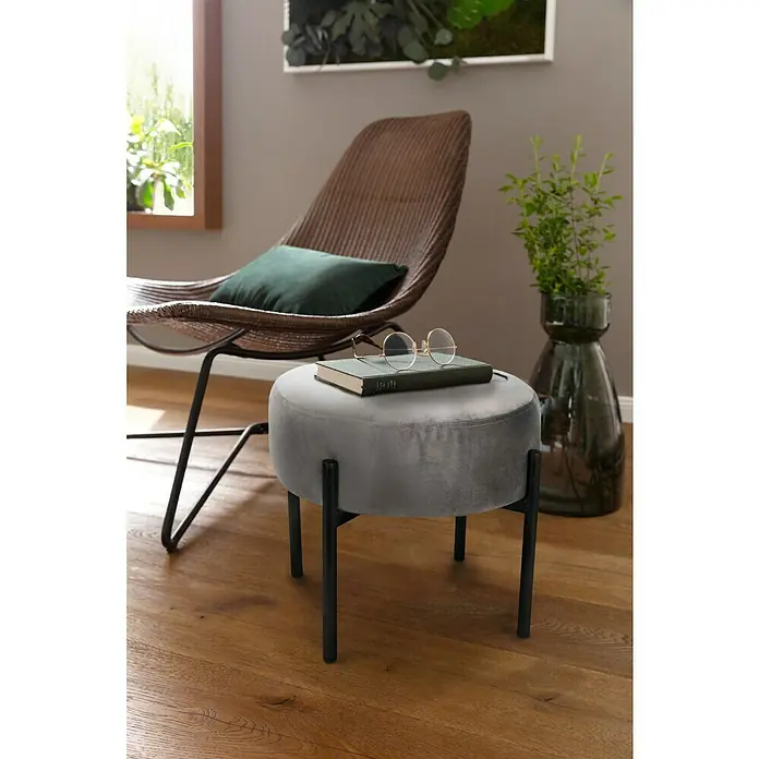 Hocker - Ø x H: 33 x 30 cm, Grau, Samt - Ein grauer, runder Samthocker mit schwarzen Beinen steht auf einem Holzboden. Auf dem Hocker liegen ein Buch und eine Brille, daneben ein Rattansessel.
