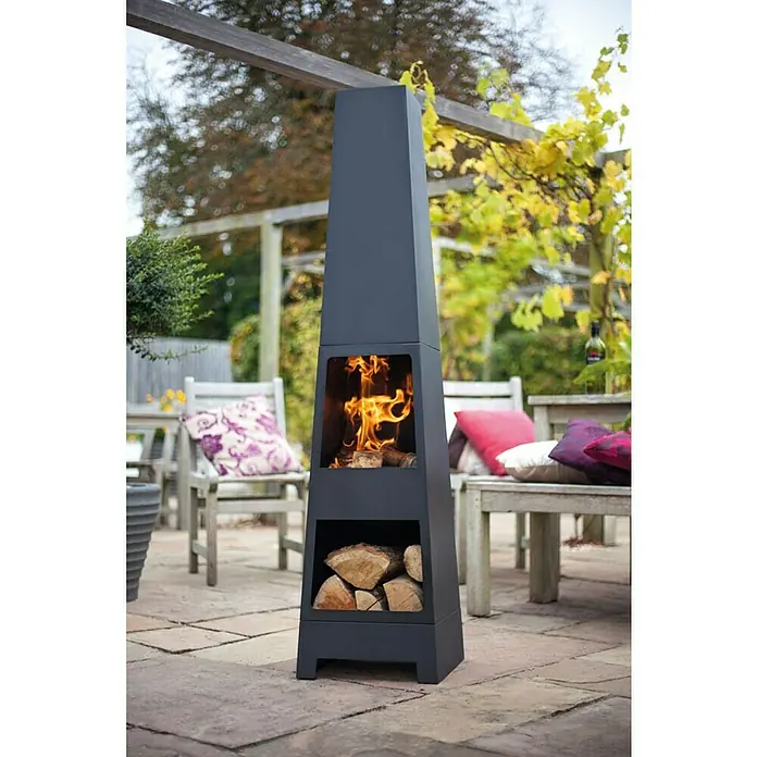 Buschbeck Chimenea para jardín Malmö - 36 x 36 x 150 cm, Acero, Recubrimiento en polvo - Una chimenea exterior oscura y moderna con troncos apilados y un fuego encendido para un ambiente cálido y acogedor al aire libre.