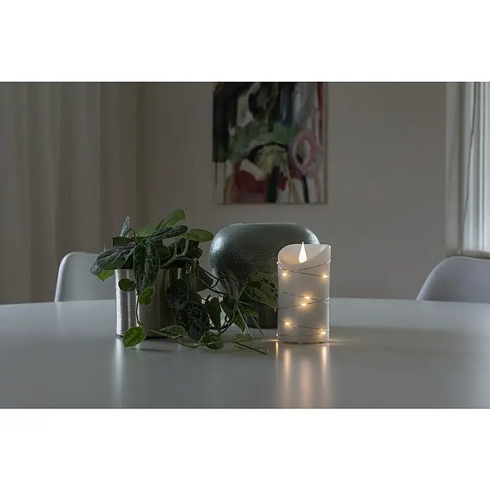 Konstsmide LED-Echtwachskerze - Ø x H: 7,5 x 13,5 cm, Creme - Tischdekoration mit einer LED-Kerze, einer Pflanze im Topf und einer grauen Vase vor einem abstrakten Gemälde.
