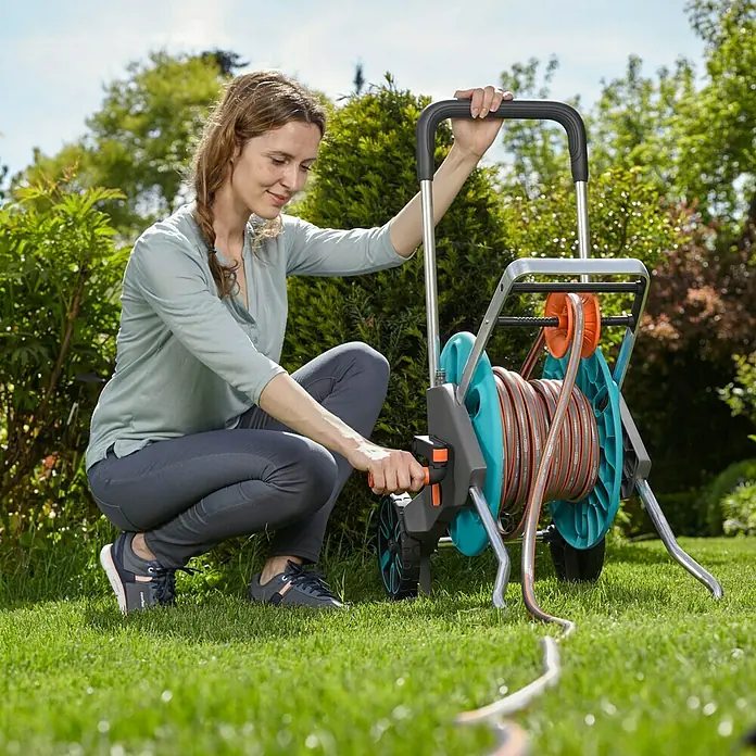 Gardena Schlauchwagen CleverRoll M EasyKunststoff/Metall, Schlauchkapazität: 60 m, 13 mm (½″) - Eine Frau bedient eine GARDENA Schlauchtrommel mit Schlauch auf einem Rasen, umgeben von Grün.