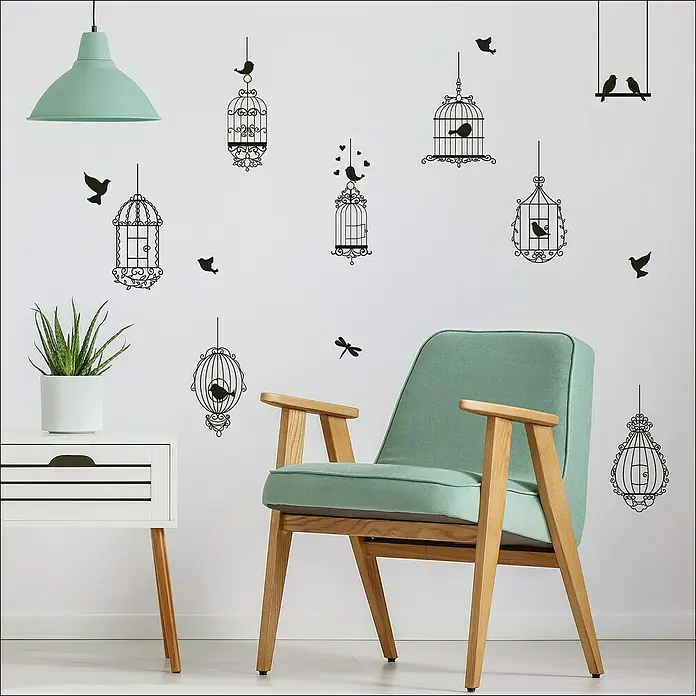 Vinilo de pared Pájaro - 48 x 68 cm, Plástico - Una silla verde menta con estructura de madera frente a una pared con motivos de jaulas de pájaros, complementada con una lámpara colgante y una mesita.
