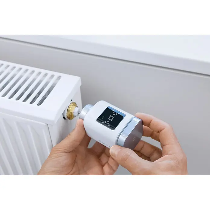 Bosch Smart Home Heizkörper-Thermostat II - M30 x 1,5 mm - Ein Heizkörperthermostat von Bosch wird von einer Hand eingestellt. Das Gerät ist weiß mit silbernen Akzenten und einem kleinen Display.