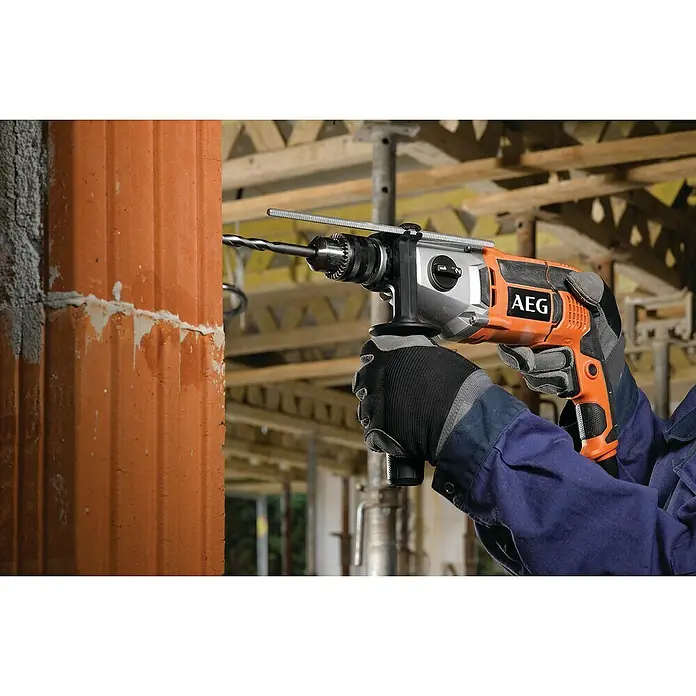 AEG Powertools Schlagbohrmaschine SB2E 1100 RV - 1.100 W, Schnellspannbohrfutter - Ein AEG-Bohrhammer wird verwendet, um Löcher in eine orangefarbene Ziegelwand zu bohren, wobei ein Baugerüst im Hintergrund zu sehen ist.