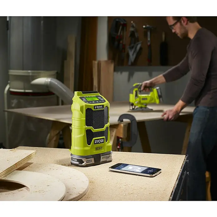 Ryobi ONE+ Radio Bluetooth de batería R18R-0 - Frecuencia: 87,5 - 108,0 kHz (FM) - Un altavoz Bluetooth Ryobi ONE+ está sobre una mesa con un teléfono inteligente al lado y un hombre usando una sierra de calar al fondo.
