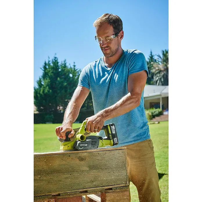 Ryobi ONE+ Cepilladora de mano de batería - 18 V, Iones de litio, Sin batería, Anchura del cepillo: 82 mm - Un hombre utiliza un cepillo eléctrico Ryobi para alisar una tabla de madera, con gafas de seguridad y trabajando al aire libre.