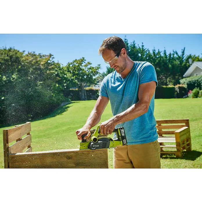 Ryobi ONE+ Cepilladora de mano de batería - 18 V, Iones de litio, Sin batería, Anchura del cepillo: 82 mm - Un hombre usa un cepillo Ryobi amarillo al aire libre sobre un palet de madera mientras usa gafas de seguridad.