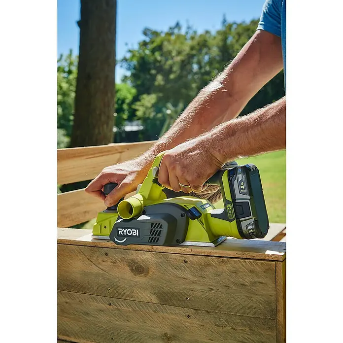 Ryobi ONE+ Cepilladora de mano de batería - 18 V, Iones de litio, Sin batería, Anchura del cepillo: 82 mm - Un hombre usa un cepillo Ryobi sobre una pieza de madera al aire libre, con un árbol y vegetación al fondo.