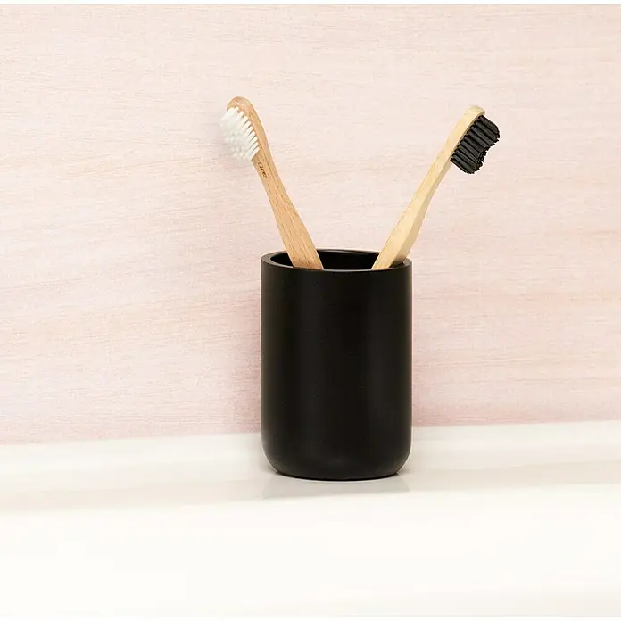 Venus Matte Black Zahnputzbecher - Polyresin, Schwarz - Zwei Holzzahnbürsten mit schwarzen und weißen Borsten stehen in einem schwarzen Becher auf einem weißen Waschbecken vor einer rosa Wand.
