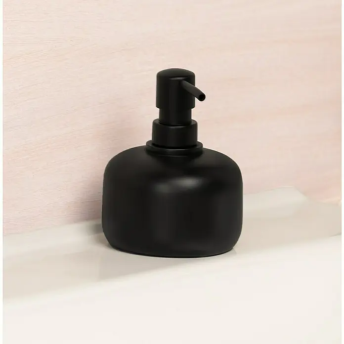 Venus Matte Black Seifenspender - Polyresin, Schwarz - Nahaufnahme einer eleganten schwarzen Seifenspenderflasche auf einer weißen Oberfläche vor einem rosafarbenen Hintergrund. Das minimalistische Design sorgt für einen modernen Touch.