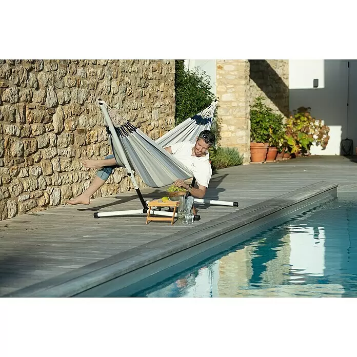 La Siesta Hängemattengestell Nautico Single Size - L x B x H: 310 x 90 x 115 cm, Stahl - Mann entspannt in einer gestreiften Hängematte auf einem Ständer am Pool, genießt Obst und Getränke.