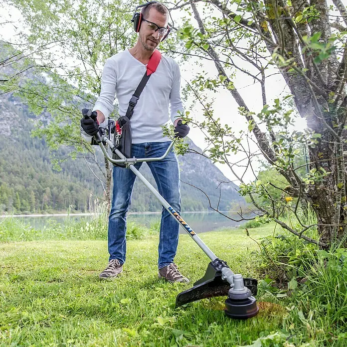 AL-KO Benzin-Motorsense BC 223 B - 700 W, Schnittbreite Faden: 41 cm, Schnittbreite Messer: 25 cm - Ein Mann benutzt einen Rasentrimmer in der Natur, umgeben von Bäumen und einem See. Der Rasentrimmer ist gut sichtbar und im Einsatz.