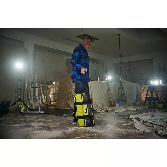 Ryobi Werkzeugtasche RSSLTB2 - Polyester - Mann zieht eine gelbe und schwarze RYOBI Werkzeugtasche auf Rädern in einem staubigen Baustellenbereich mit hellen Arbeitsleuchten.
