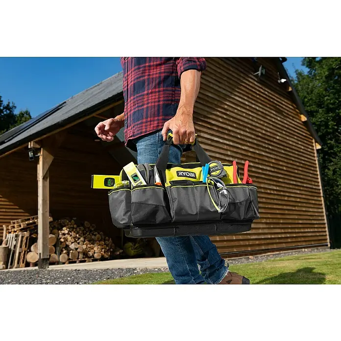 Ryobi Werkzeugtasche RSSMTB1 M - Polyester, Größe: M - Ein Handwerker trägt eine Ryobi Werkzeugtasche, gefüllt mit Werkzeugen, vor einem Holzhaus.
