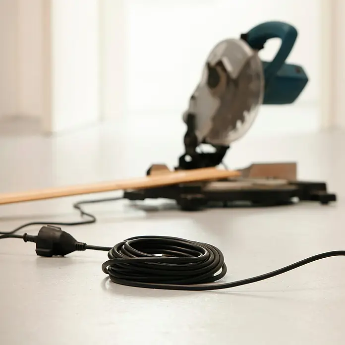 Voltomat Verlängerungskabel - 10 m, Schwarz, H05VV-F - Eine Kreissäge steht auf dem Boden, mit einem aufgerollten Kabel im Vordergrund. Perfekt für Holzarbeiten und Heimwerkerprojekte.