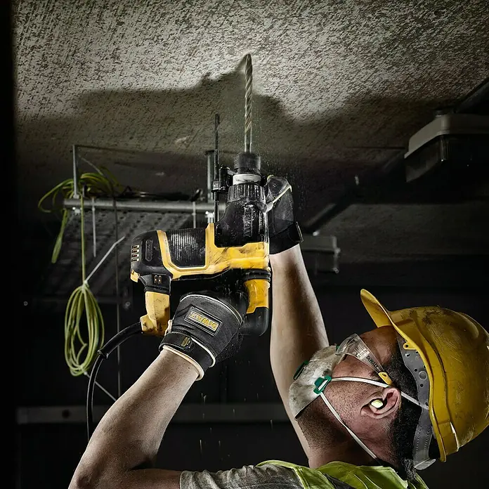 DeWalt Broca para hormigón y mampostería DT9553-QZ - 1 ud., Diámetro de la broca: 12 mm - Un trabajador de la construcción perfora agujeros en un techo con un taladro amarillo, mientras usa gafas protectoras y un casco.