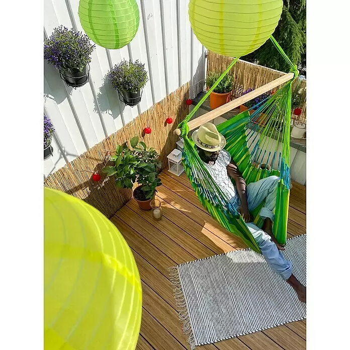 La Siesta Hängesessel Domingo Kingsize - Lime - Eine Person entspannt in einer lebhaften grün-blau gestreiften Hängematte mit Holzspreizstab auf einem sonnigen Balkon mit Holzdeck.