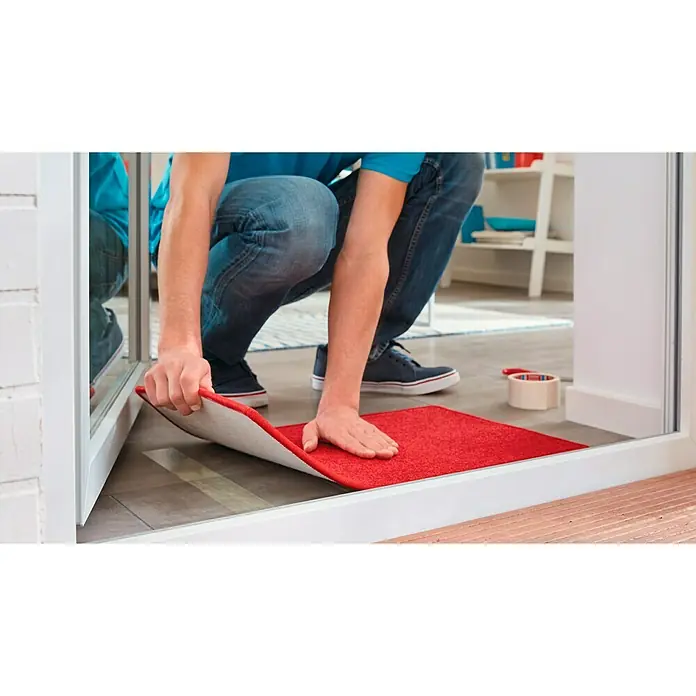 Tesa Cinta de doble cara para moquetas - 50 mm x 10 m - Un hombre está colocando un felpudo rojo. La alfombra tiene un respaldo antideslizante y es ideal para entradas.