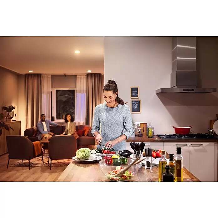 Philips Hue Smart-LED-Lampen Set White - E27, Dimmbar, Warmweiß, 1.055 lm, 9,5 W, 2 Stk. - Eine Frau schneidet Gemüse in einer hell beleuchteten Küche, während zwei Personen in einem warm beleuchteten Wohnzimmer entspannen.