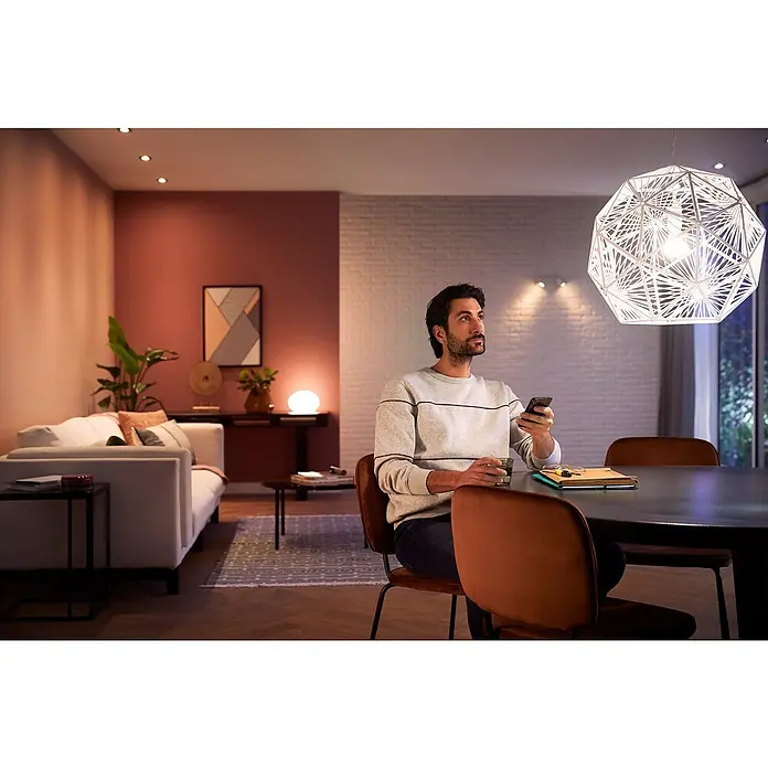 Philips Hue LED-Lampe WhiteE27, Dimmbar, Warmweiß, 1.055 lm, 9,5 W - Ein Mann sitzt am Tisch und steuert die intelligente Beleuchtung im Wohnzimmer mit seinem Handy. Moderne Beleuchtungslösung für Ihr Zuhause.