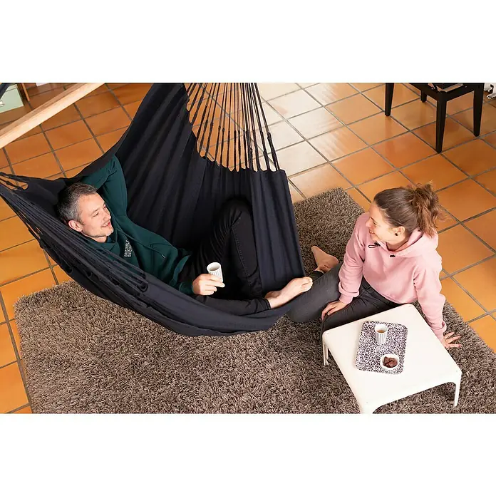 La Siesta Hängesessel Habana Comfort - L x B x H: 180 x 115 x 160 cm, Onyx - Ein Mann entspannt in einer schwarzen Hängematte mit einer Tasse, während eine Frau daneben sitzt und Kaffee auf einem Tisch steht.