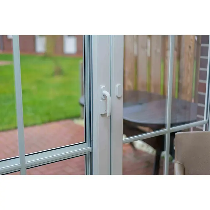 Homematic IP Fenstersensor HmIP-SRH - Weiß, 16 x 32 x 143 mm, Batteriebetrieben - Nahaufnahme einer weißen Terrassentür mit Griff und Blick auf eine Terrasse mit Tisch und Stuhl. Die Tür trennt Innen- und Außenbereich.