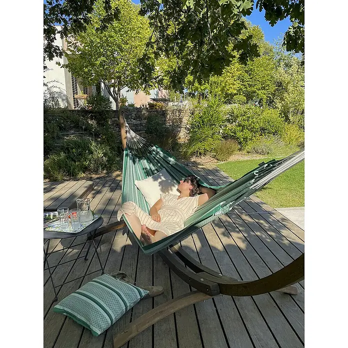 La Siesta Hängematte Modesta - L x B: 350 x 160 cm, Agave, Double - Eine Frau entspannt in einer grünen Hängematte auf einer Holzterrasse, neben einem Tisch mit Getränken. Sommerliche Entspannung im Freien.