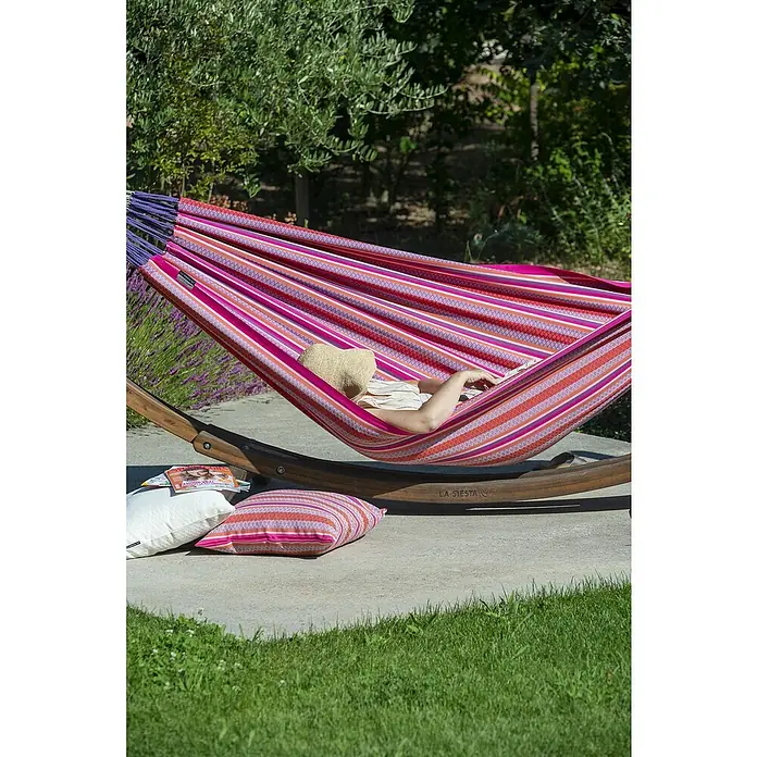La Siesta Hängematte Modesta - L x B: 350 x 160 cm, Flamingo, Double - Eine Person entspannt in einer bunten Hängematte von La Siesta im Garten. Die Hängematte hängt zwischen zwei Holzständern und bietet Komfort.