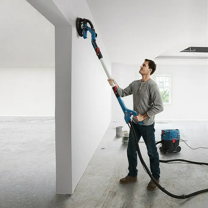 Bosch Professional Wand- & Deckenschleifer GTR 55-225 - 550 W, Durchmesser Schleifteller: 225 mm - Ein Mann benutzt einen Bosch Trockenbauschleifer mit Staubsauger zum Schleifen einer Wand. Das Schleifgerät ist blau und hat einen langen Griff.